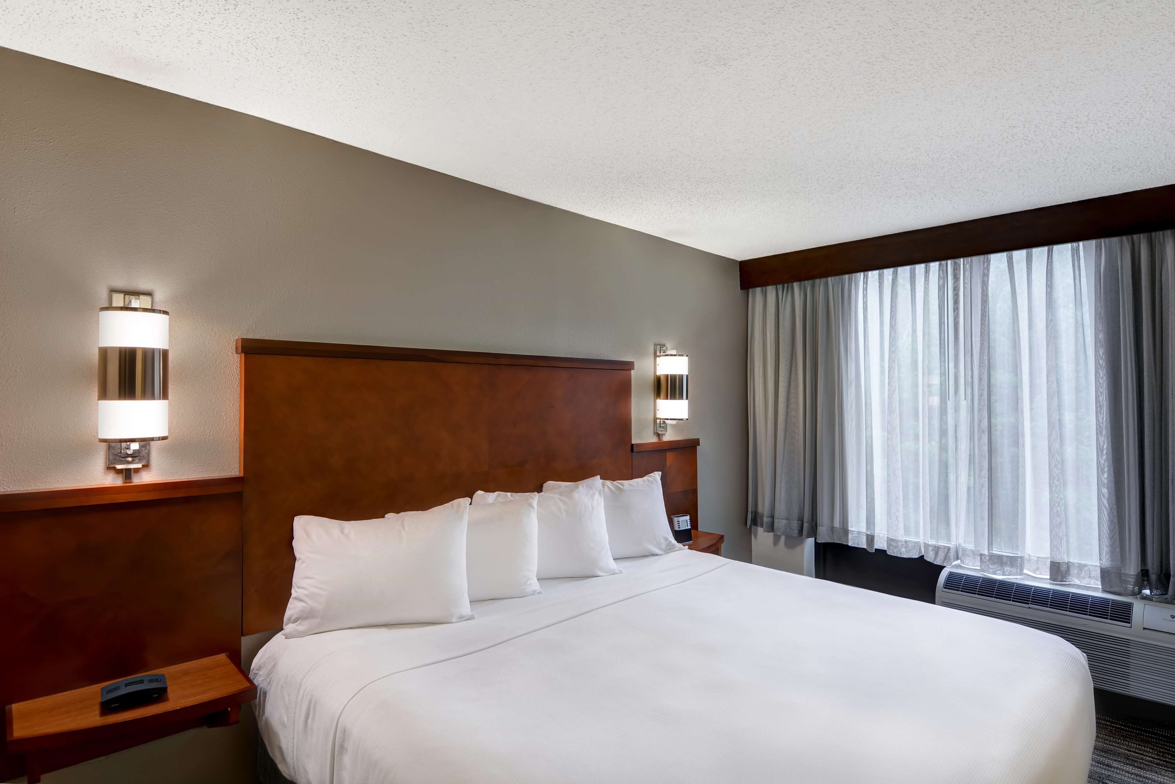 A hotel room with a large bed, two bedside tables, a nightstand, a wall-mounted lamp, a window with curtains, and an air conditioning unit.