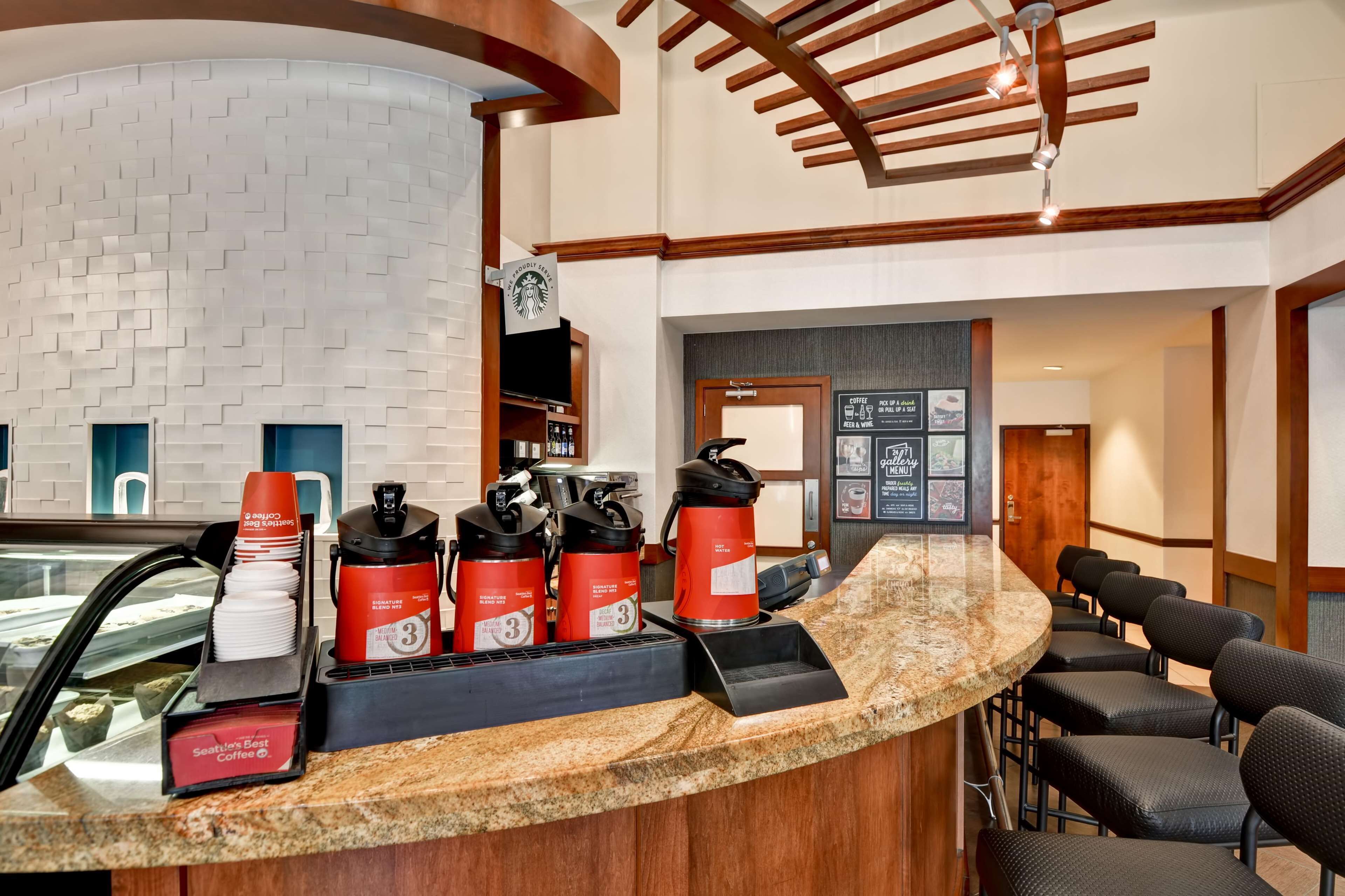 Zona de recepción de un hotel con un mostrador curvo de granito, una máquina de café Starbucks y exhibición de tazas con la marca Seattle's Best Coffee.