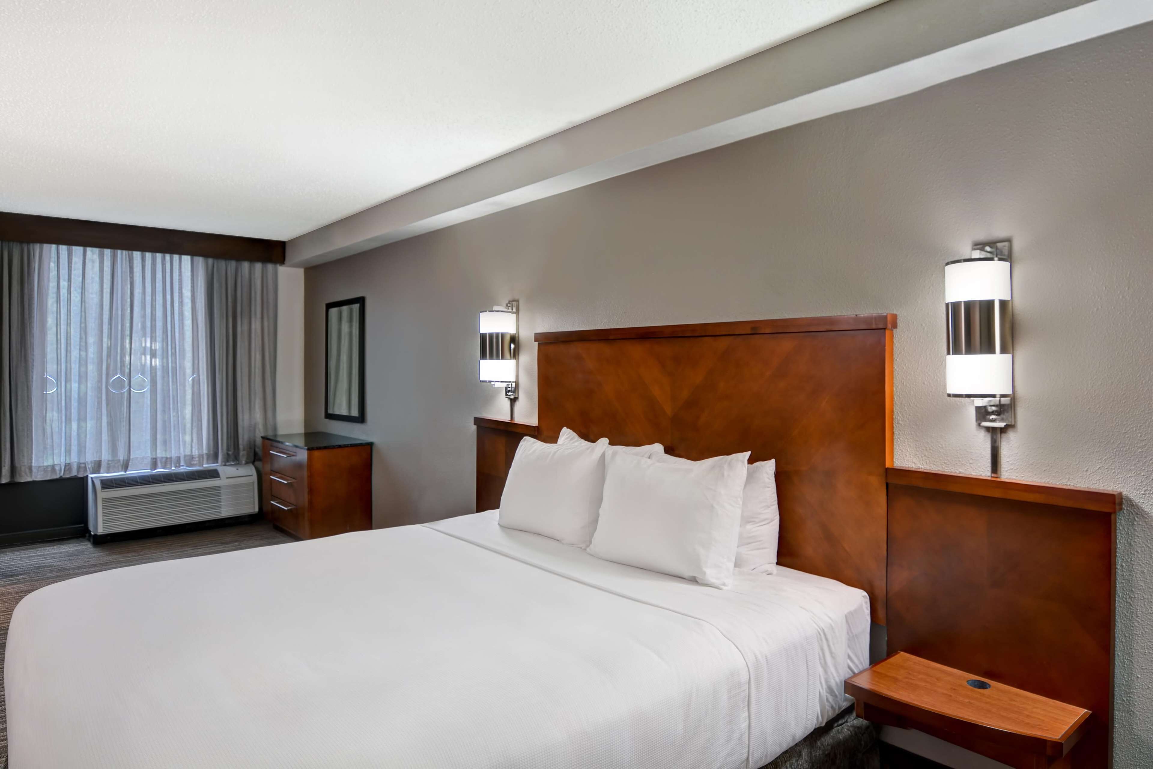 A hotel room with a large bed, a wooden headboard, two bedside tables with lamps, a window with curtains, and a wall-mounted air conditioning unit.