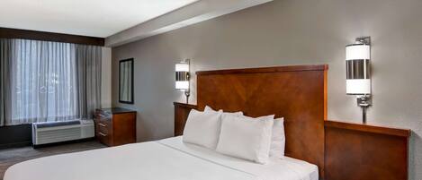 A hotel room with a large bed, a wooden headboard, two bedside tables with lamps, a window with curtains, and a wall-mounted air conditioning unit.
