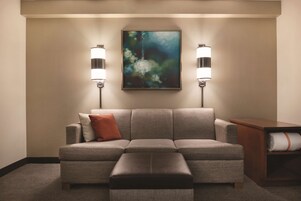 Una sala de estar en tonos neutros con un sofá gris, un reposapiés marrón y un cuadro en la pared.