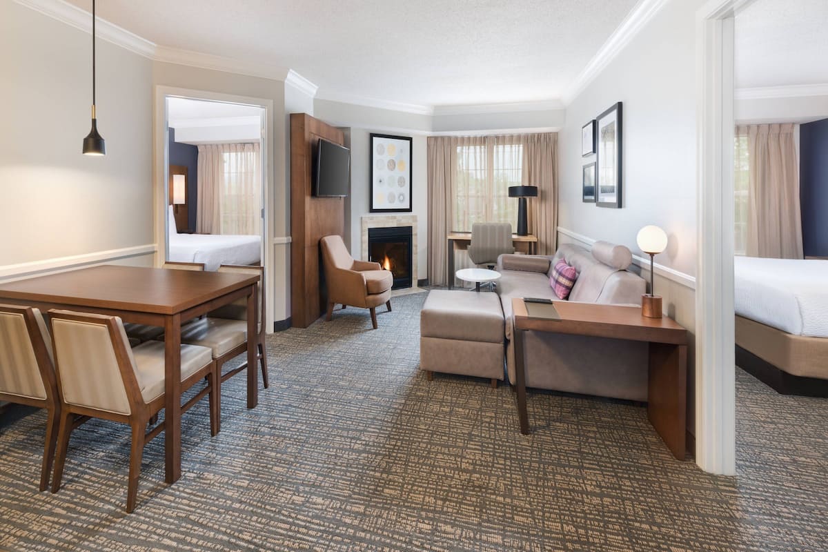 A hotel room with a dining table, two chairs, a sofa, a small desk, a lamp, a TV, and a fireplace.