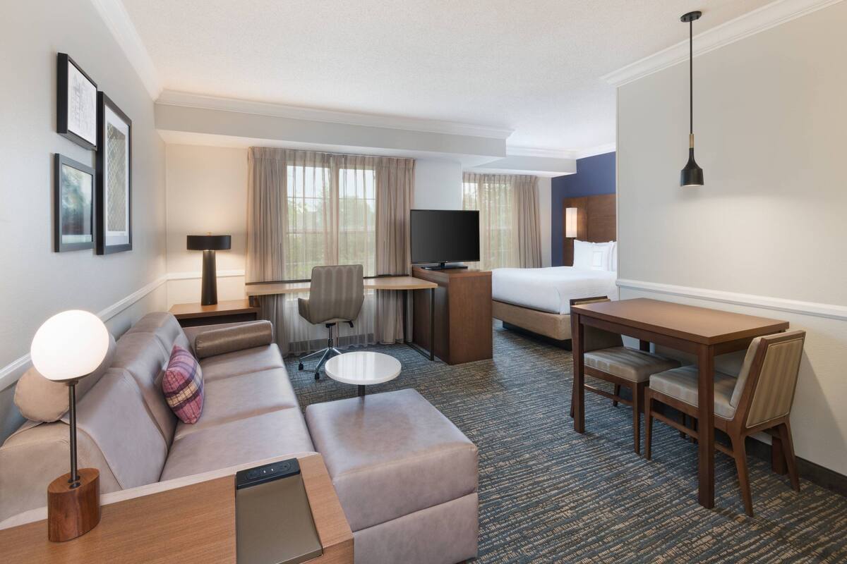 A hotel room with a sofa, a small round table, a bed, a desk with a chair, and a TV.