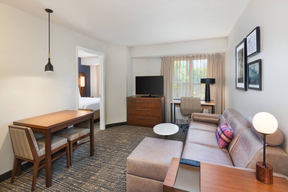 A hotel room with a dining table, sofa, TV, and a desk with a lamp.
