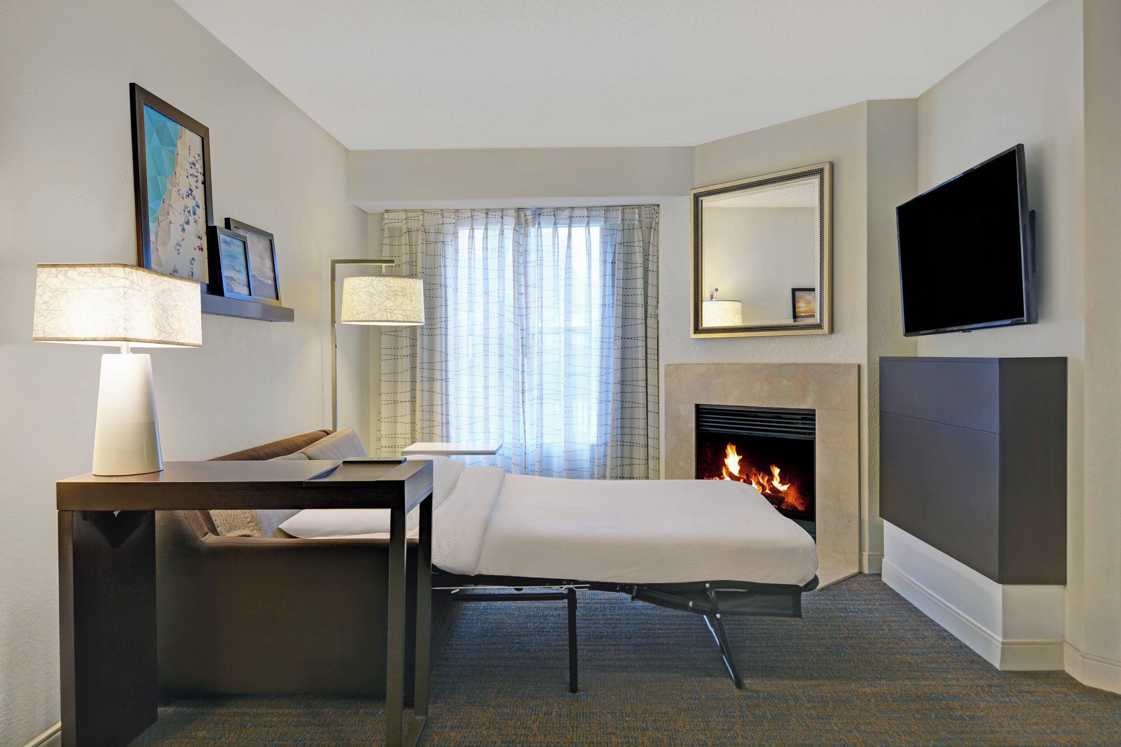 A hotel room with a bed, bedside table, lamp, fireplace, TV, and artwork on the wall.