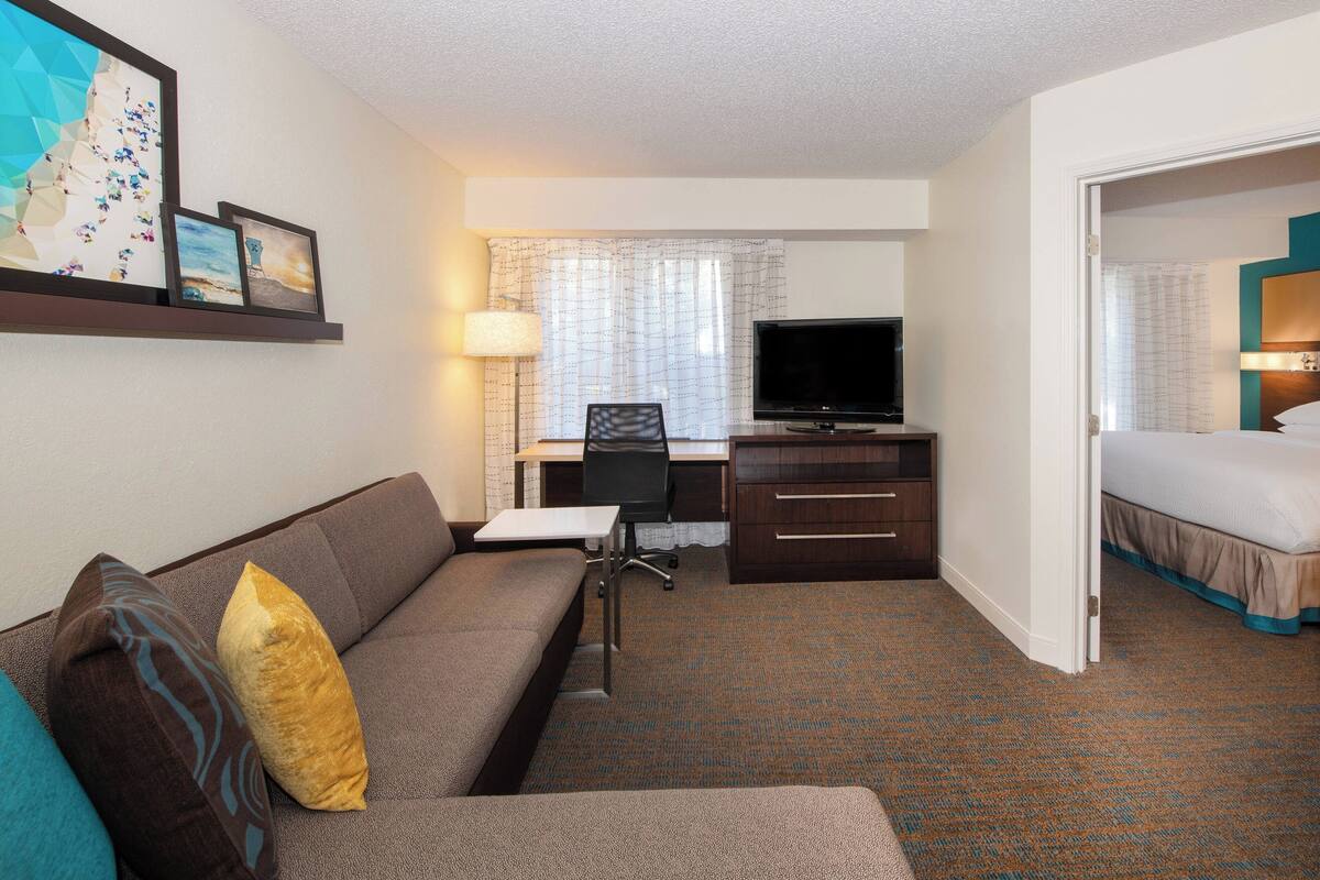 A hotel room with a sofa, a desk with a chair, a television, a bed, and a door leading to another room.