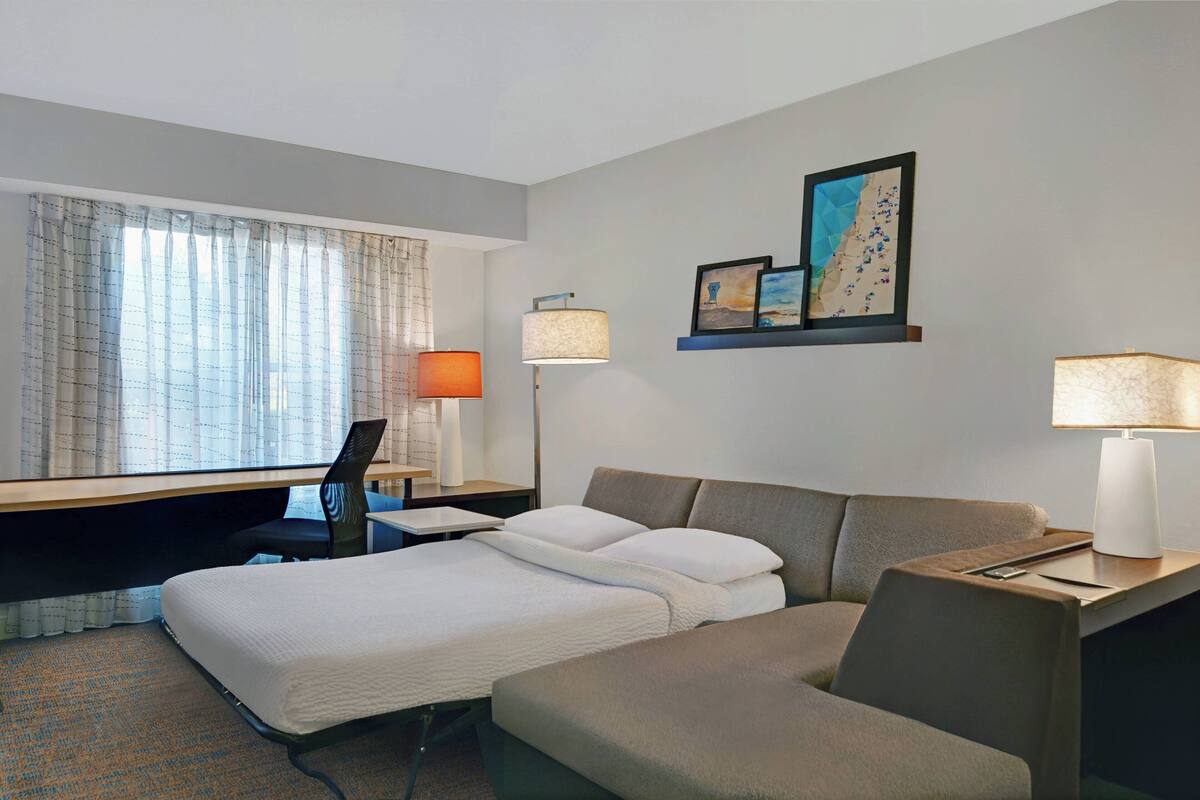 A hotel room with a bed, a desk, a chair, a lamp, and a wall-mounted shelf with pictures.