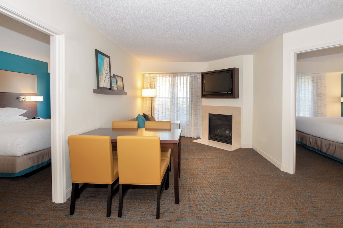A hotel room with a bed, a desk with chairs, a television, a fireplace, and a window with curtains.