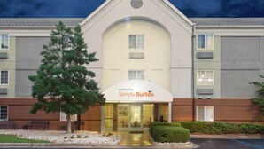 Un hotel con diseño moderno, que destaca por su entrada con techo curvo y un letrero que dice "Sonesta Simply Suites". Hay bancos y un césped bien cuidado frente al edificio.