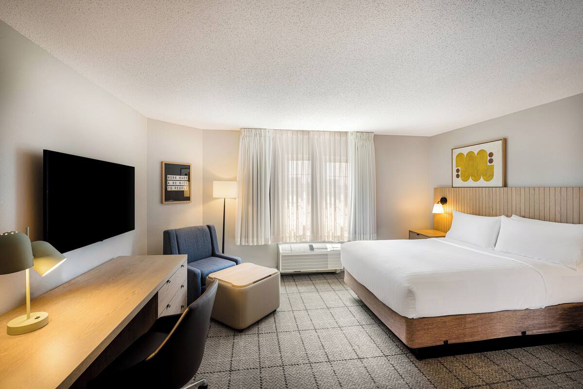 A hotel room with a bed, a desk with a lamp, a chair, a TV, a window with curtains, and a wall-mounted picture.
