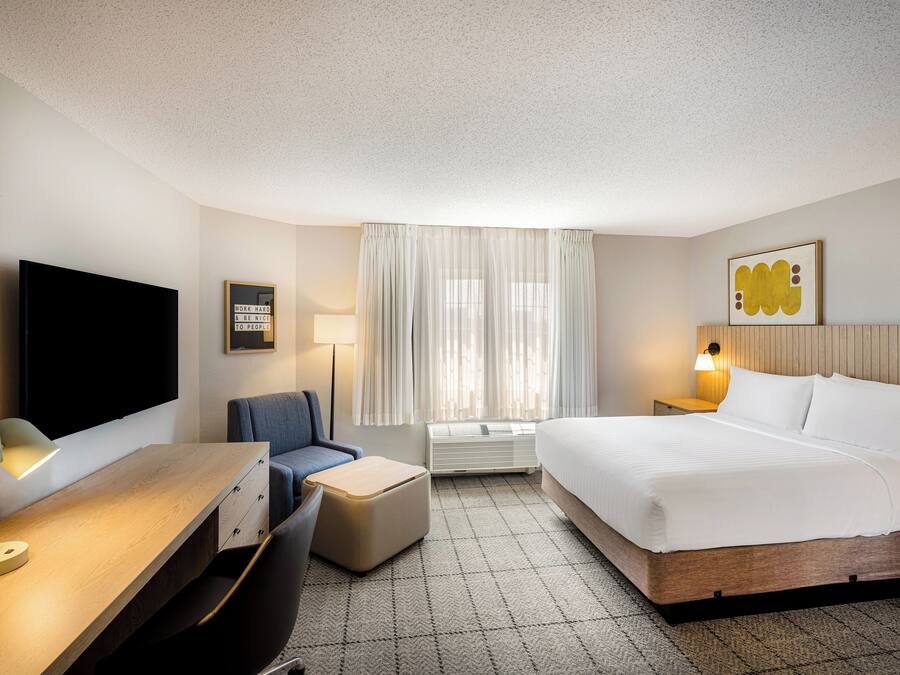 Una habitación de hotel con una cama, un escritorio con lámpara, una silla, un televisor, una ventana con cortinas y un cuadro colgado en la pared.