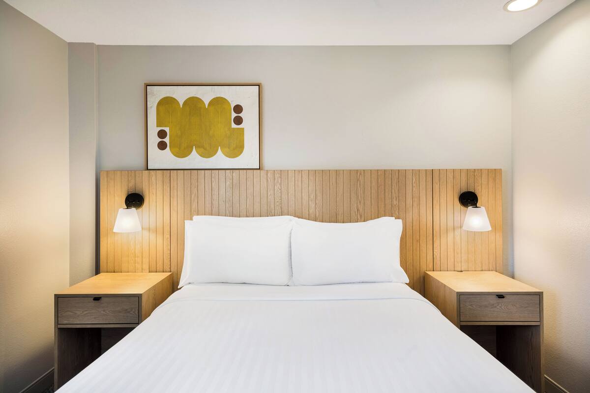 A hotel room with a wooden headboard, two bedside tables with lamps, a large bed with white linens, and a framed abstract artwork on the wall.
