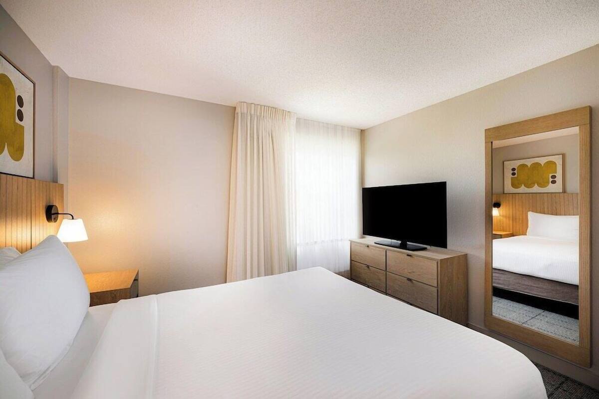 A hotel room with a large bed, a television, a bedside table with a lamp, a mirror, and a window with curtains.