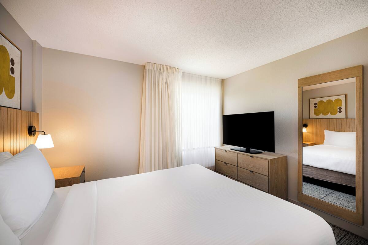 A hotel room with a large bed, a television on a wooden dresser, a mirror, and a wall-mounted lamp.