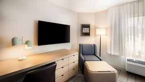Habitación de hotel con televisión de pantalla plana, escritorio con cajones, una silla, un pequeño reposapiés, una lámpara y un ventanal grande con cortinas translúcidas.