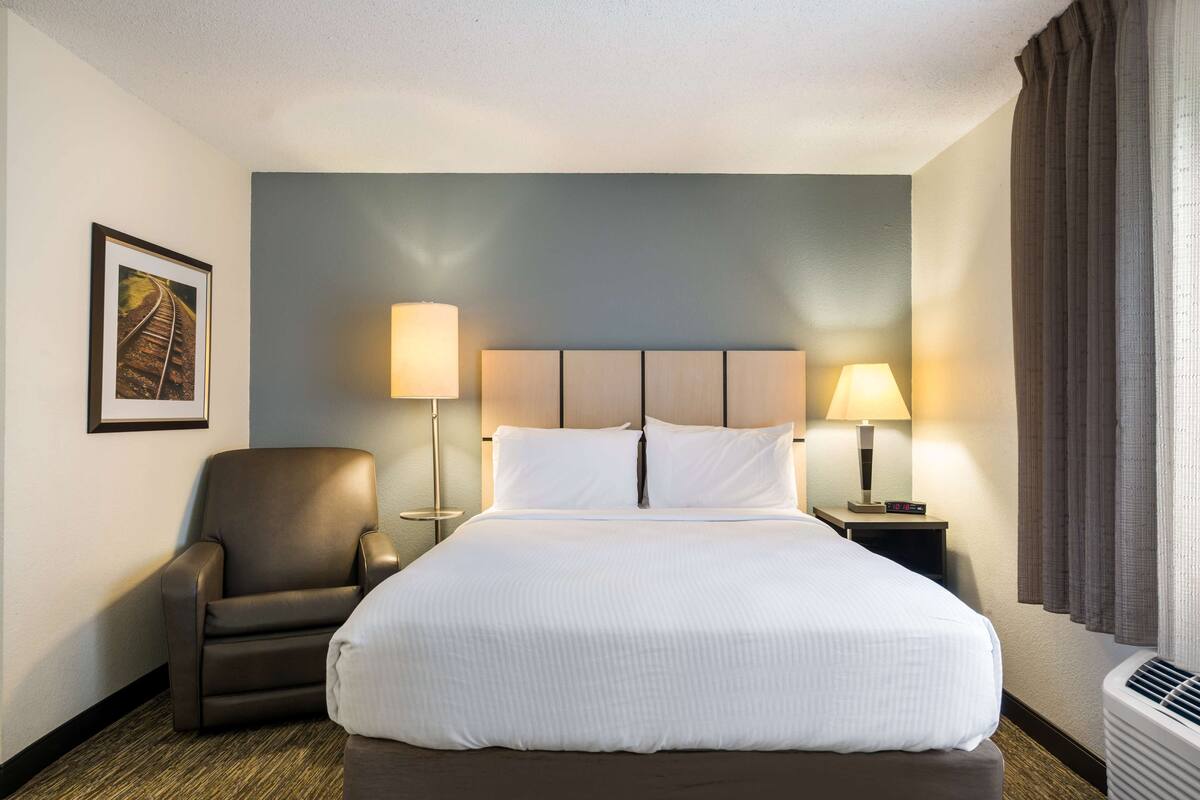 Une chambre d’hôtel avec un grand lit, deux lampes de chevet, une chaise, un tableau encadré au mur et une fenêtre avec des rideaux.