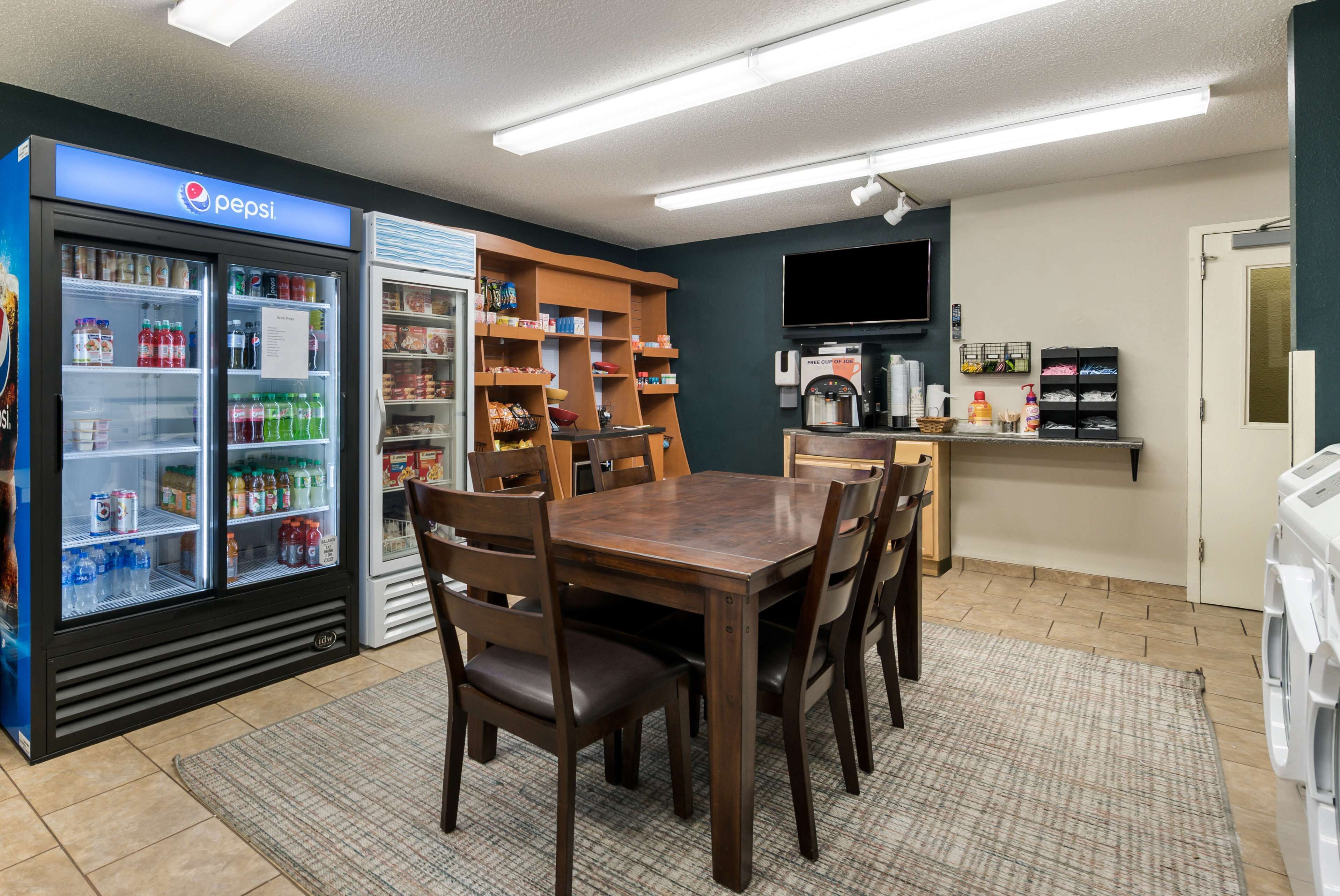 A compact hotel room with a dining area featuring a wooden table and chairs, a refrigerator stocked with beverages, a coffee machine, and a TV mounted on the wall.