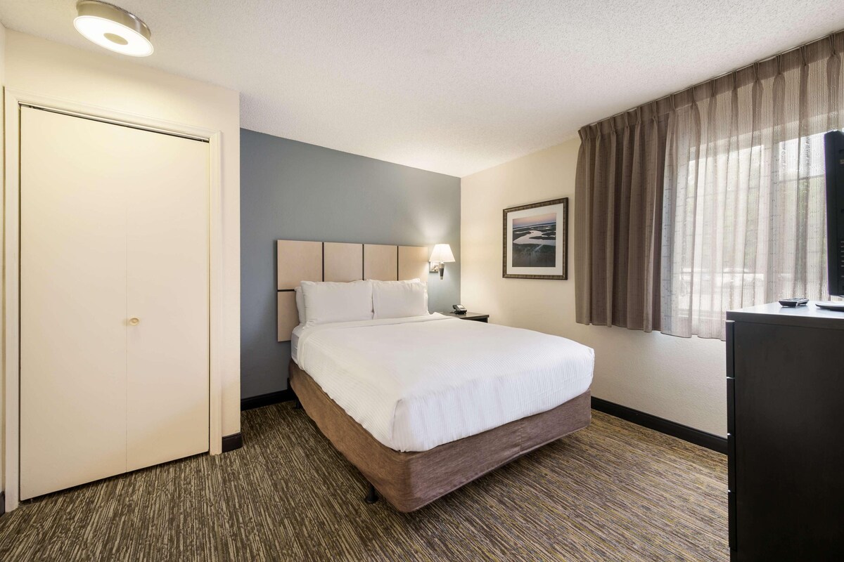 Une chambre d’hôtel avec un lit, une table de chevet, une lampe, une porte, une fenêtre avec des rideaux et un tableau au mur.