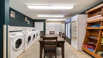 A laundry room with a table and chairs, a refrigerator, and a shelf with snacks.