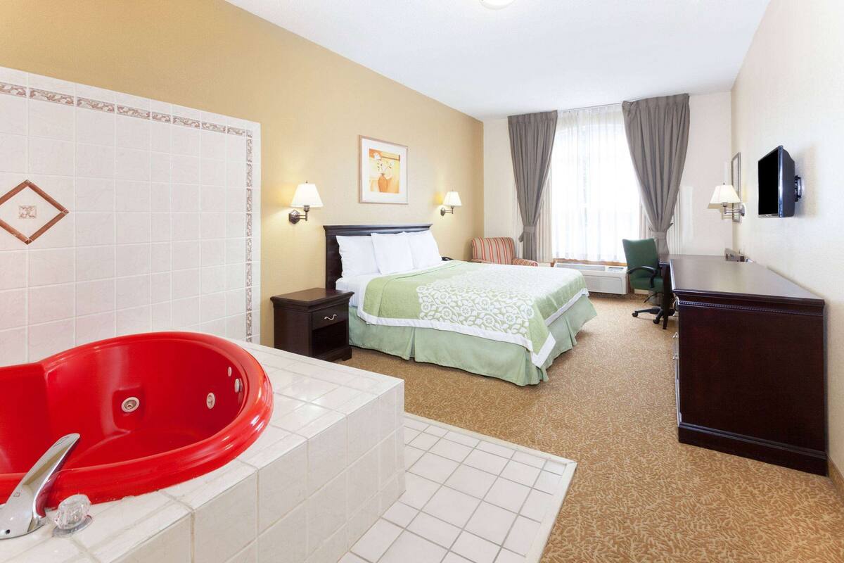 A hotel room with a red bathtub, a bed with green bedding, a desk, and a chair.
