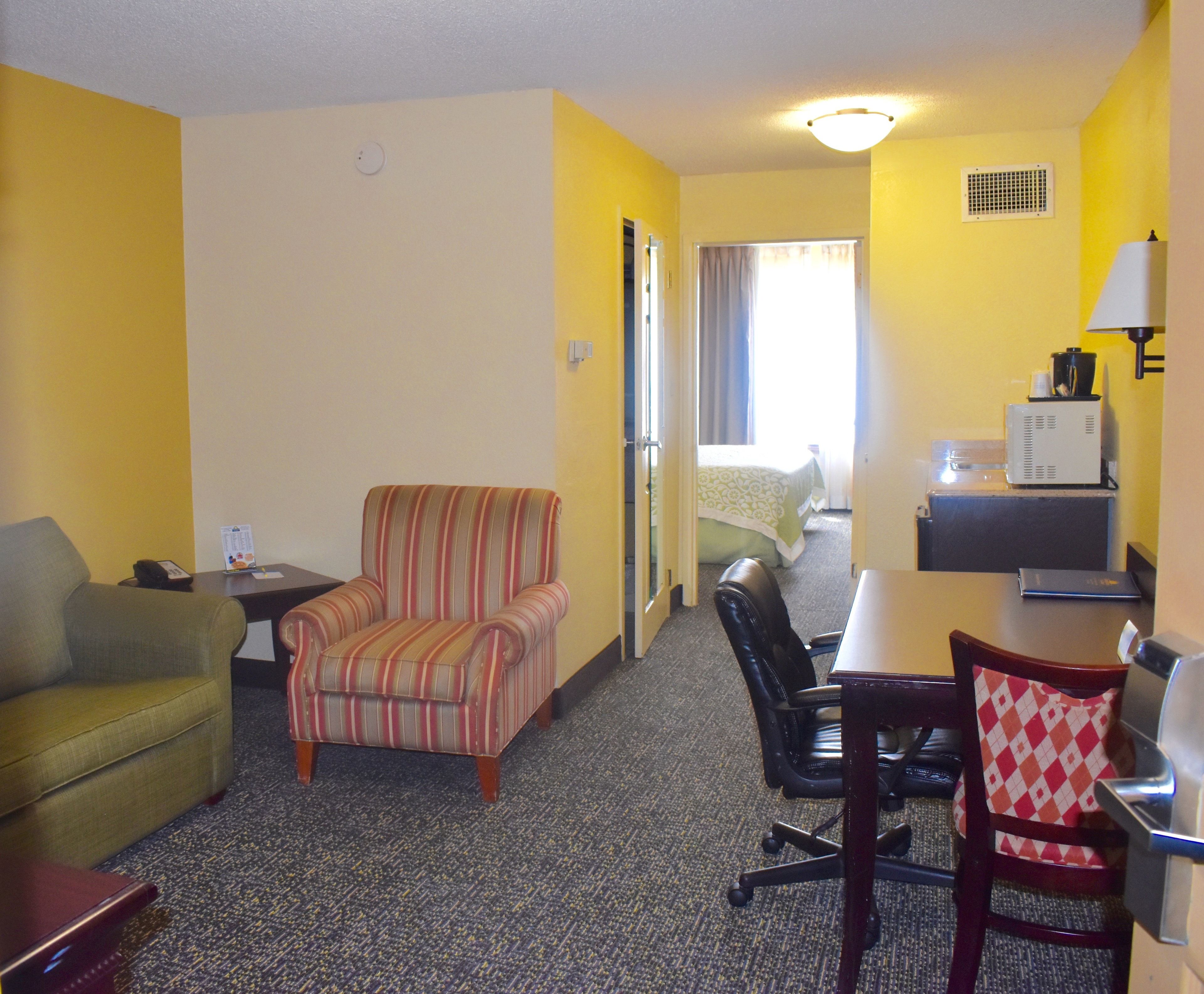 A hotel room with a striped armchair, a green sofa, a dining table with a chair, a small desk with a lamp, and a kitchenette area.