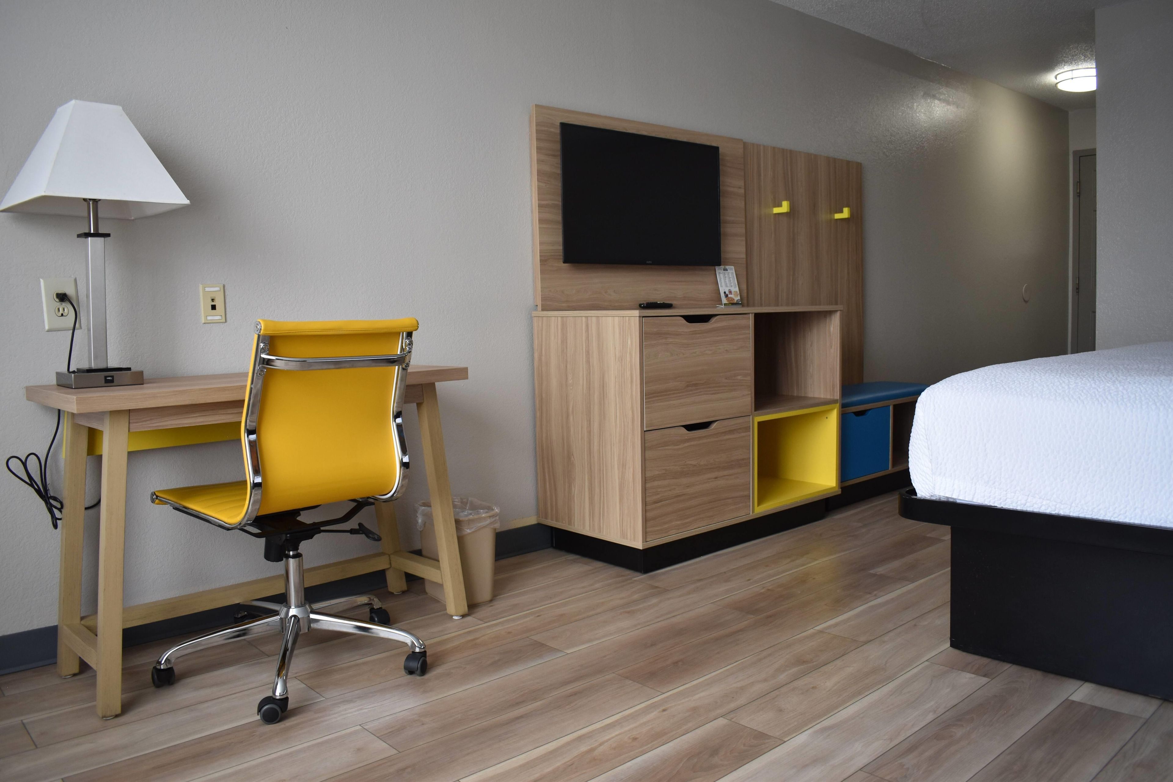A hotel room with a wooden desk, a yellow office chair, a bed, a TV on the wall, and a small cabinet with drawers.