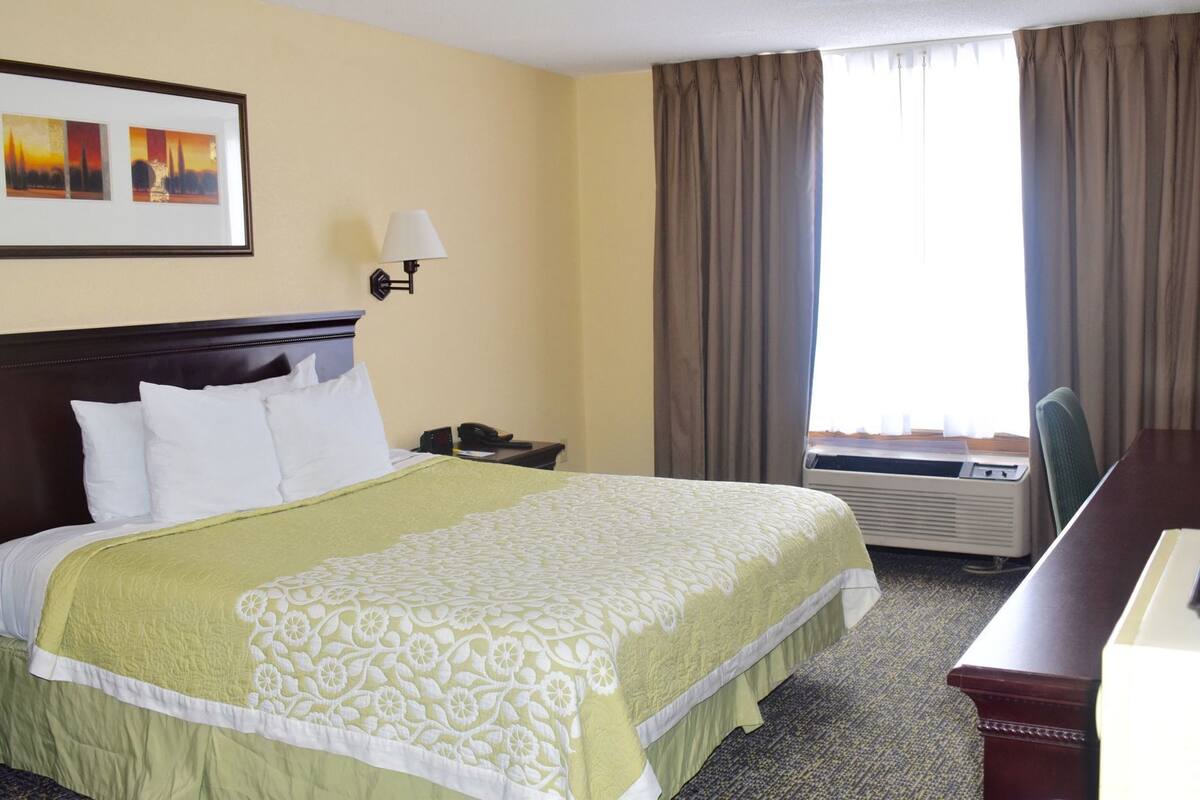A hotel room with a bed, a nightstand, a lamp, a desk, and an air conditioning unit.