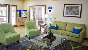 A lobby with a green armchair, a green sofa, a glass coffee table with a vase of white flowers, a framed picture on the wall, and a bulletin board with a map. - Days Inn & Suites by Wyndham Tucker/Northlake (Tucker)