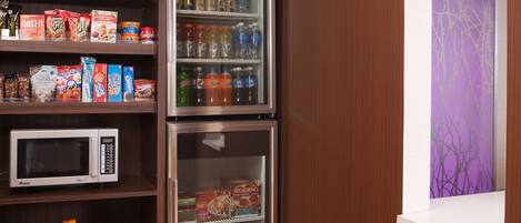 A kitchenette with a microwave, a refrigerator stocked with snacks and beverages, and a window with a purple view.