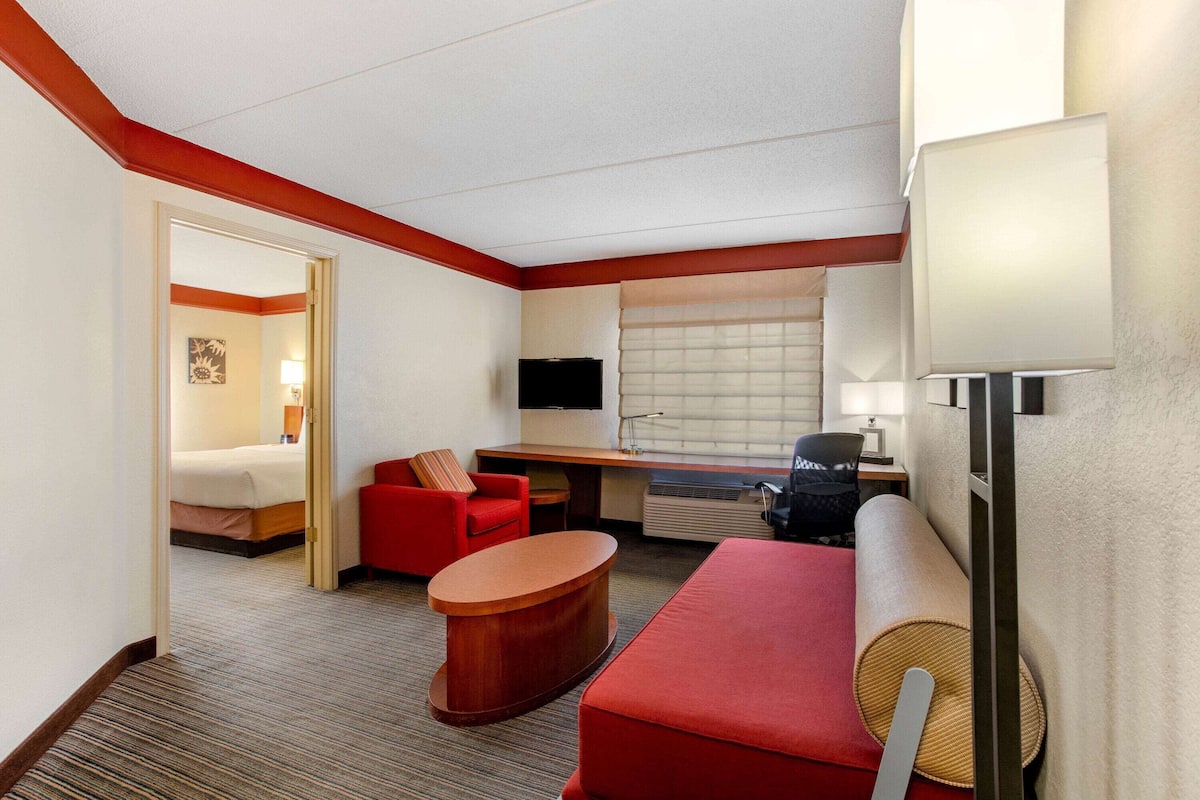 A hotel room with a bed, a red sofa, a desk with a computer, a TV, and a window with blinds.
