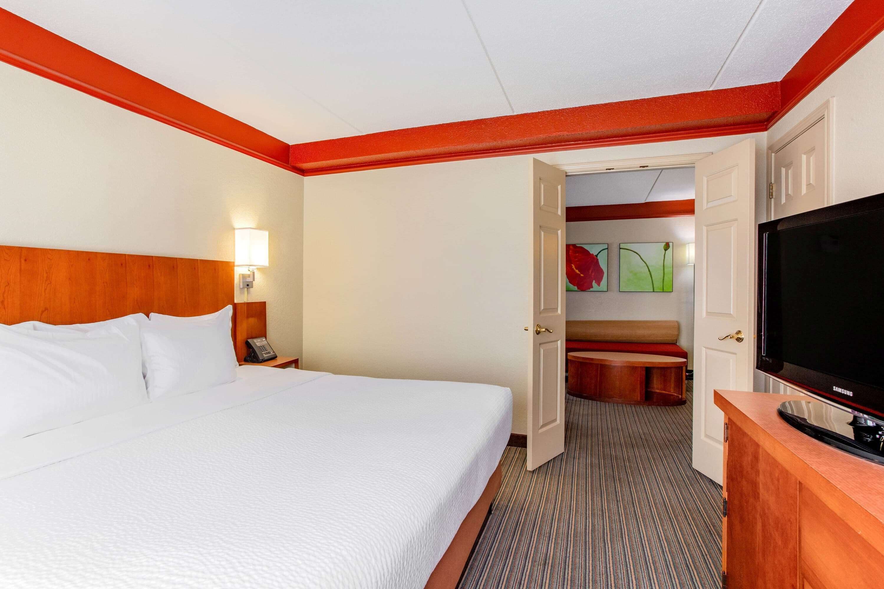 Habitación de hotel con una cama grande, un televisor sobre un soporte de madera y una puerta que da a una zona de estar.