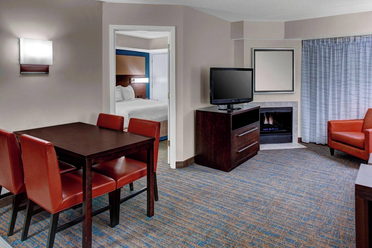 A hotel room with a dining table, red chairs, a television, a fireplace, and a large window with curtains.