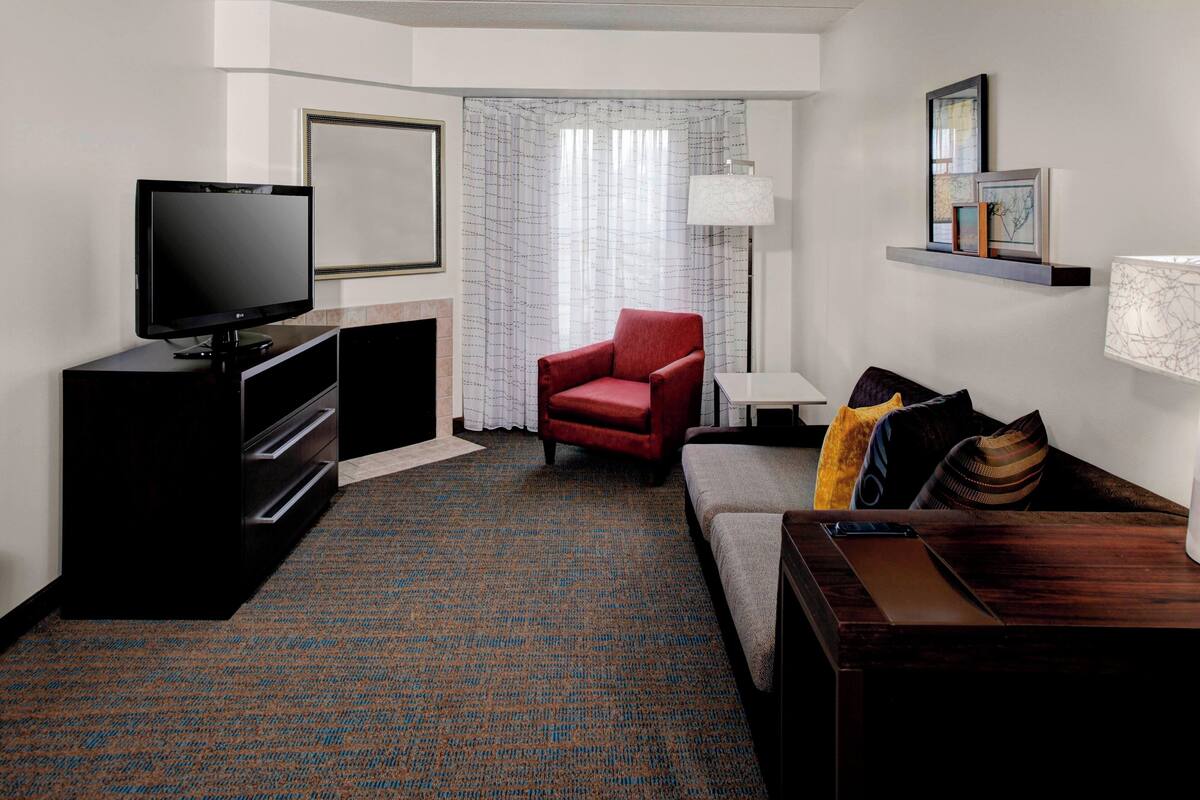 A hotel room with a flat-screen TV, a red armchair, a sofa with cushions, a small table, and a lamp.