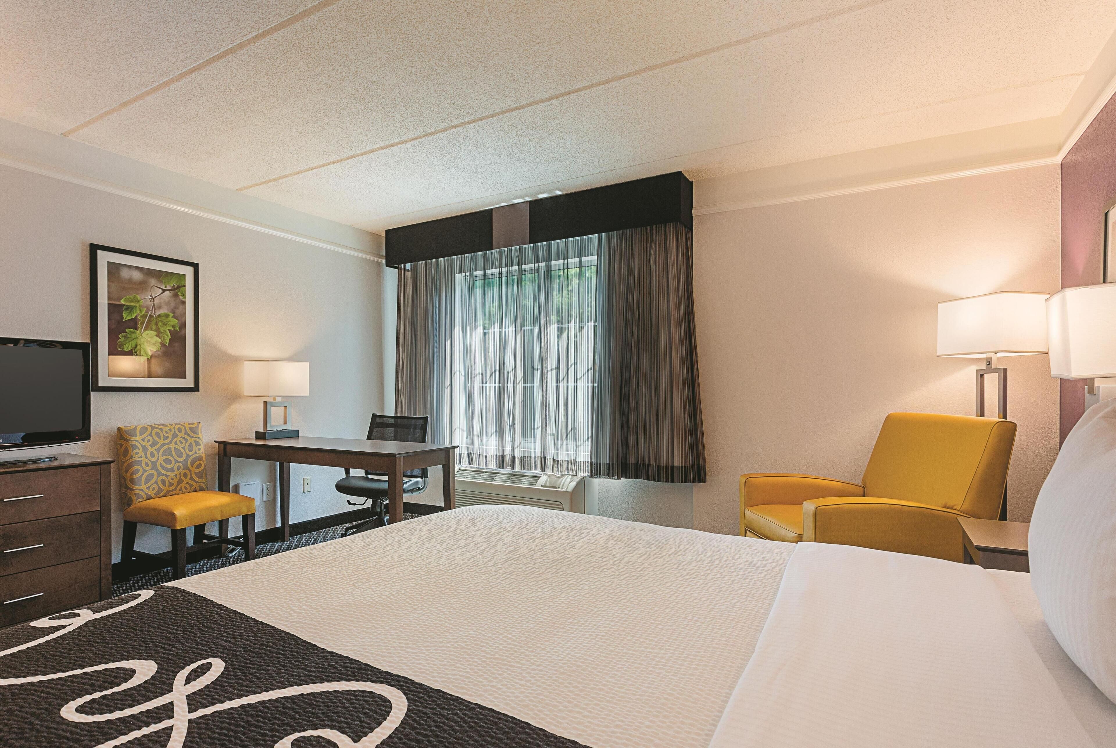 Habitación de hotel con una cama grande, un escritorio, dos sillas, un televisor y una ventana con cortinas.