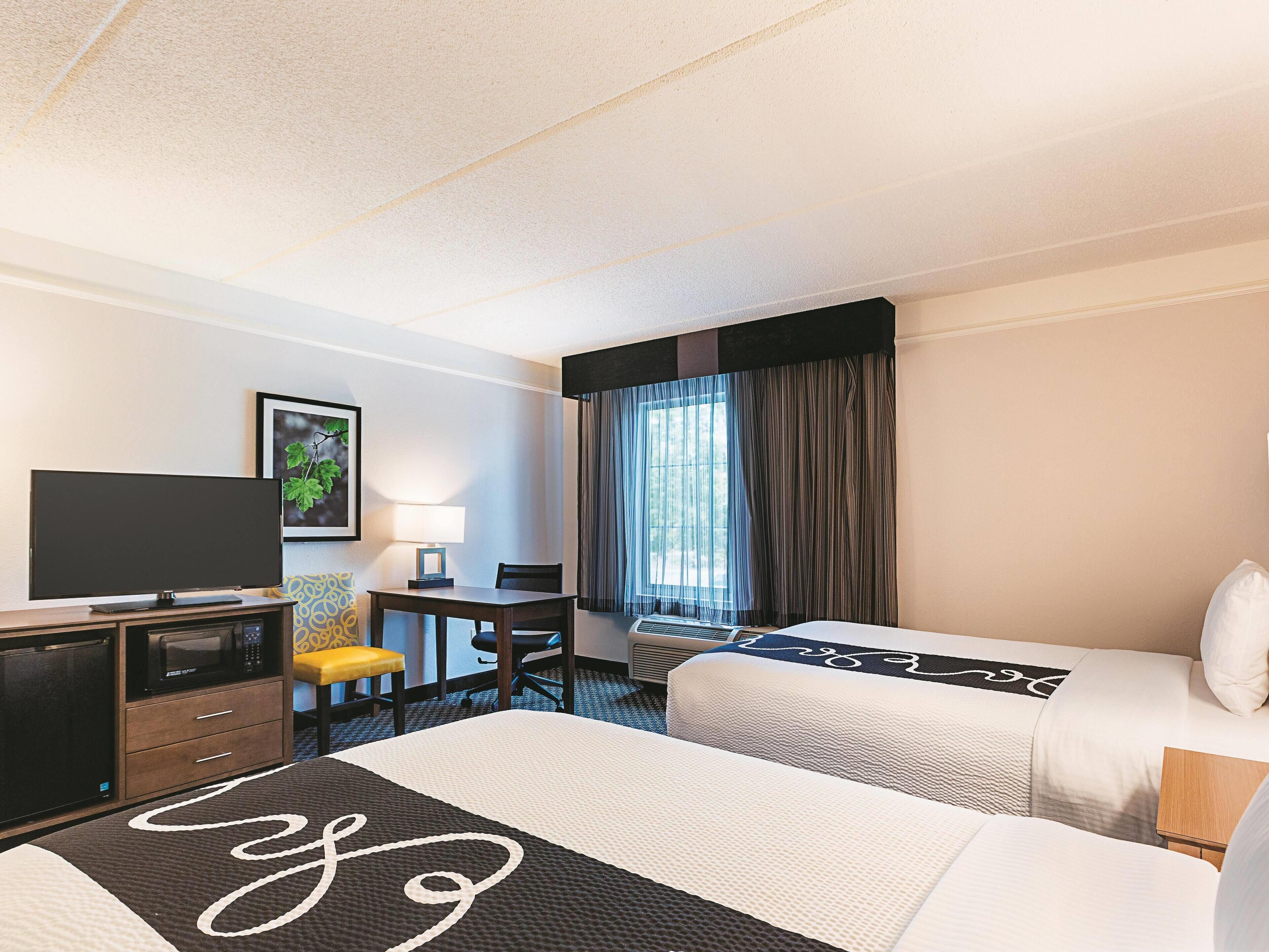 A hotel room with two beds, a desk, a television, and a window with curtains.