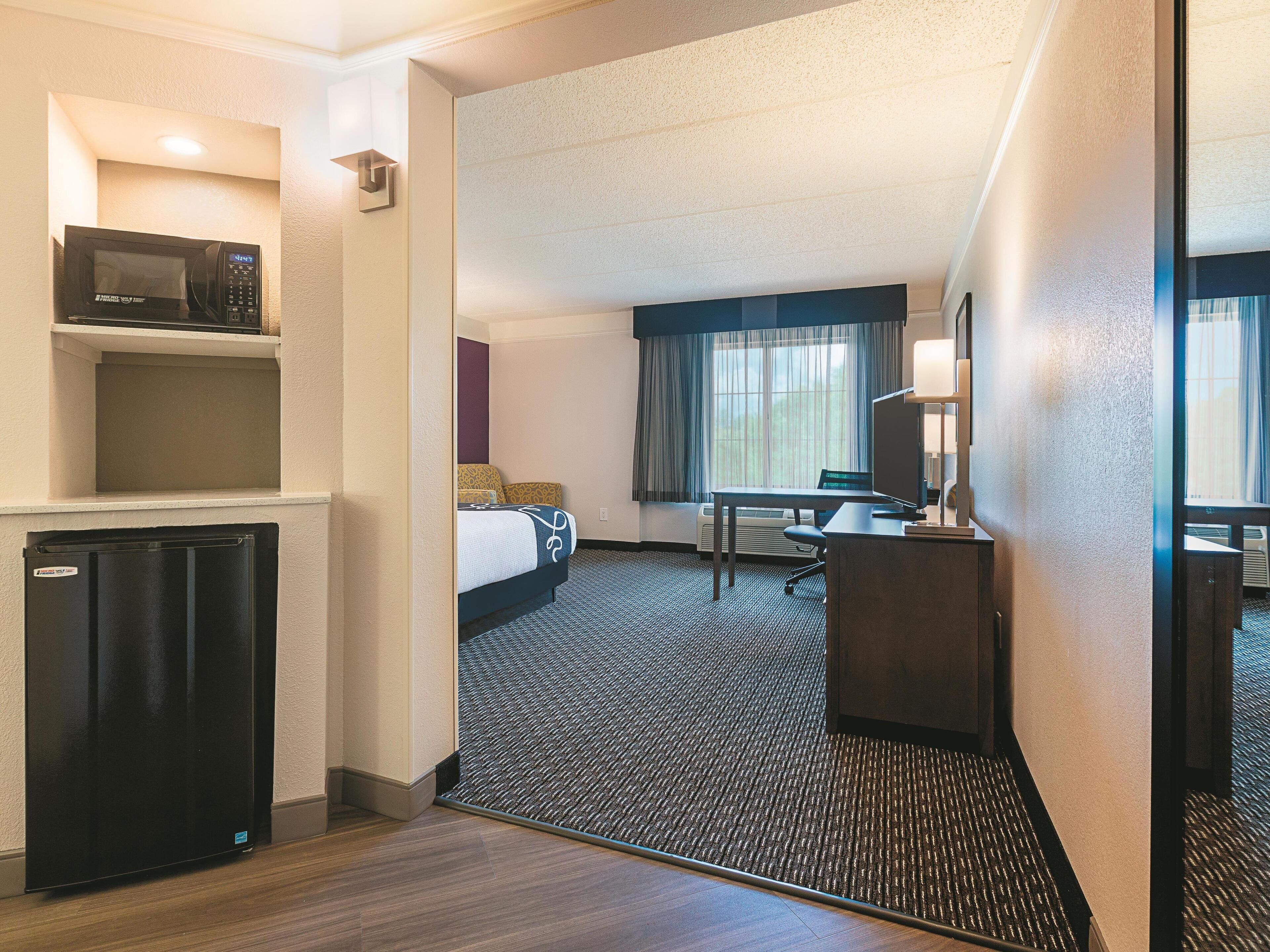 Habitación de hotel con cama, escritorio, silla, microondas, refrigerador y un amplio ventanal con cortinas.