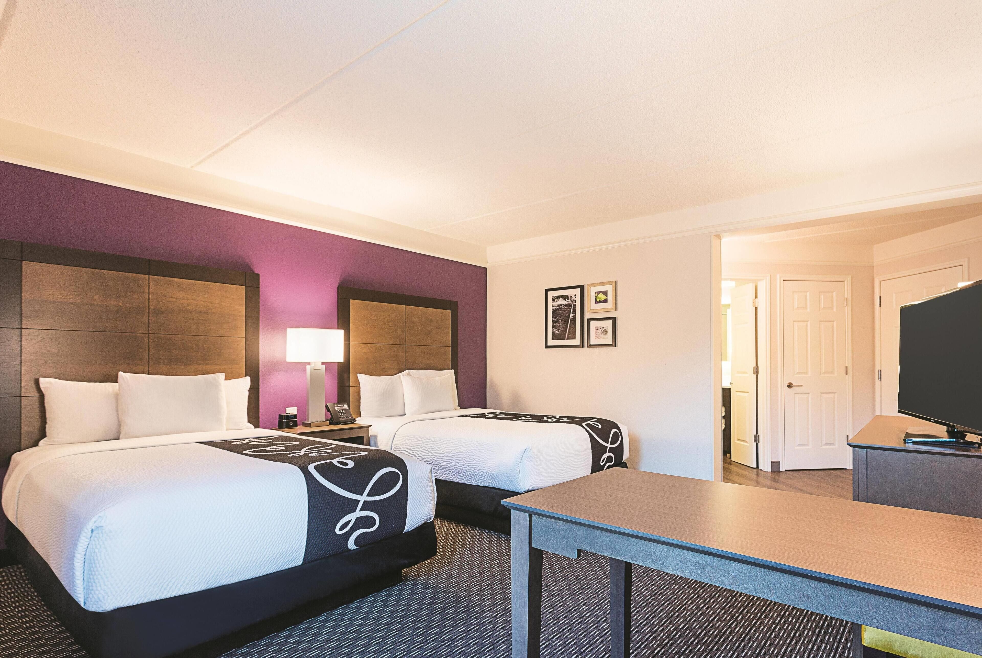 A hotel room with two beds, a desk, a TV, and a door leading to another room.