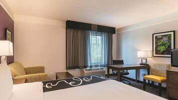 Quarto de hotel com uma cama grande, uma escrivaninha, uma cadeira, uma mesinha, uma televisão e uma janela com cortinas.