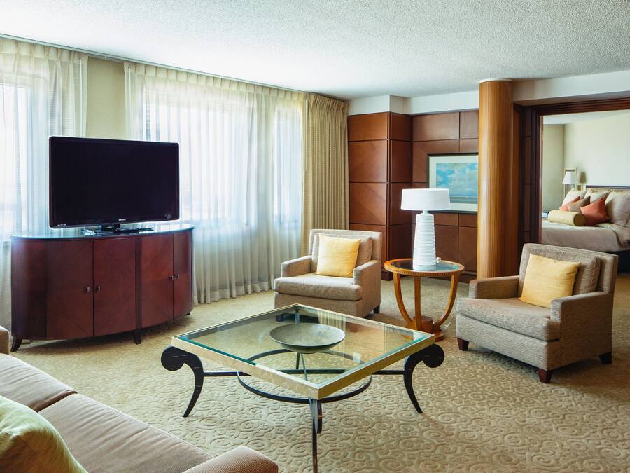 Habitación de hotel con televisor de pantalla plana, dos sillones, una mesa de centro de vidrio, un armario de madera y un amplio ventanal con cortinas.
