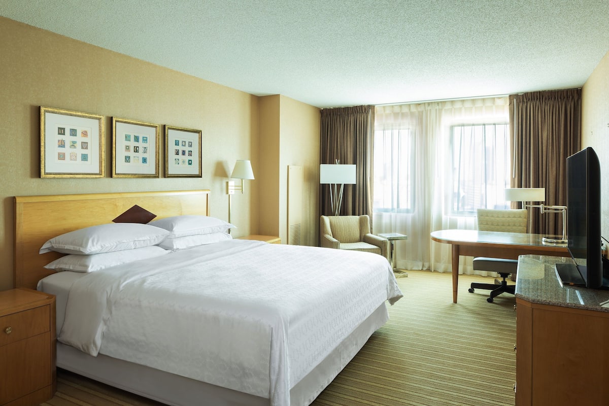 A hotel room with a large bed, a desk with a chair, a TV, and framed pictures on the wall.