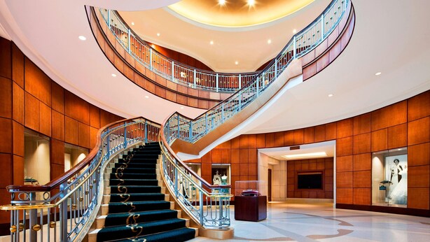Eine imposante, geschwungene Treppe mit Glastrennwand, die zu einer Lobby mit hohen Decken, Holzvertäfelung und einer zentralen Statue führt.