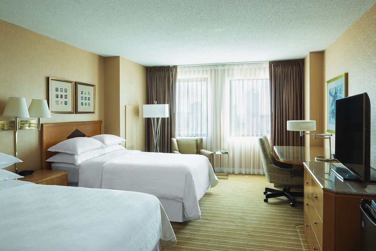 A hotel room with two beds, a desk, a chair, a TV, and a lamp.