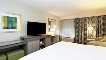 A hotel room with a large bed, a desk with a flat-screen TV, a chair, a small table, a lamp, and a window with curtains.