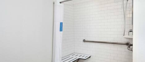 A bathroom with white tiled walls, a shower with a handheld showerhead, a grab bar, and a bench.