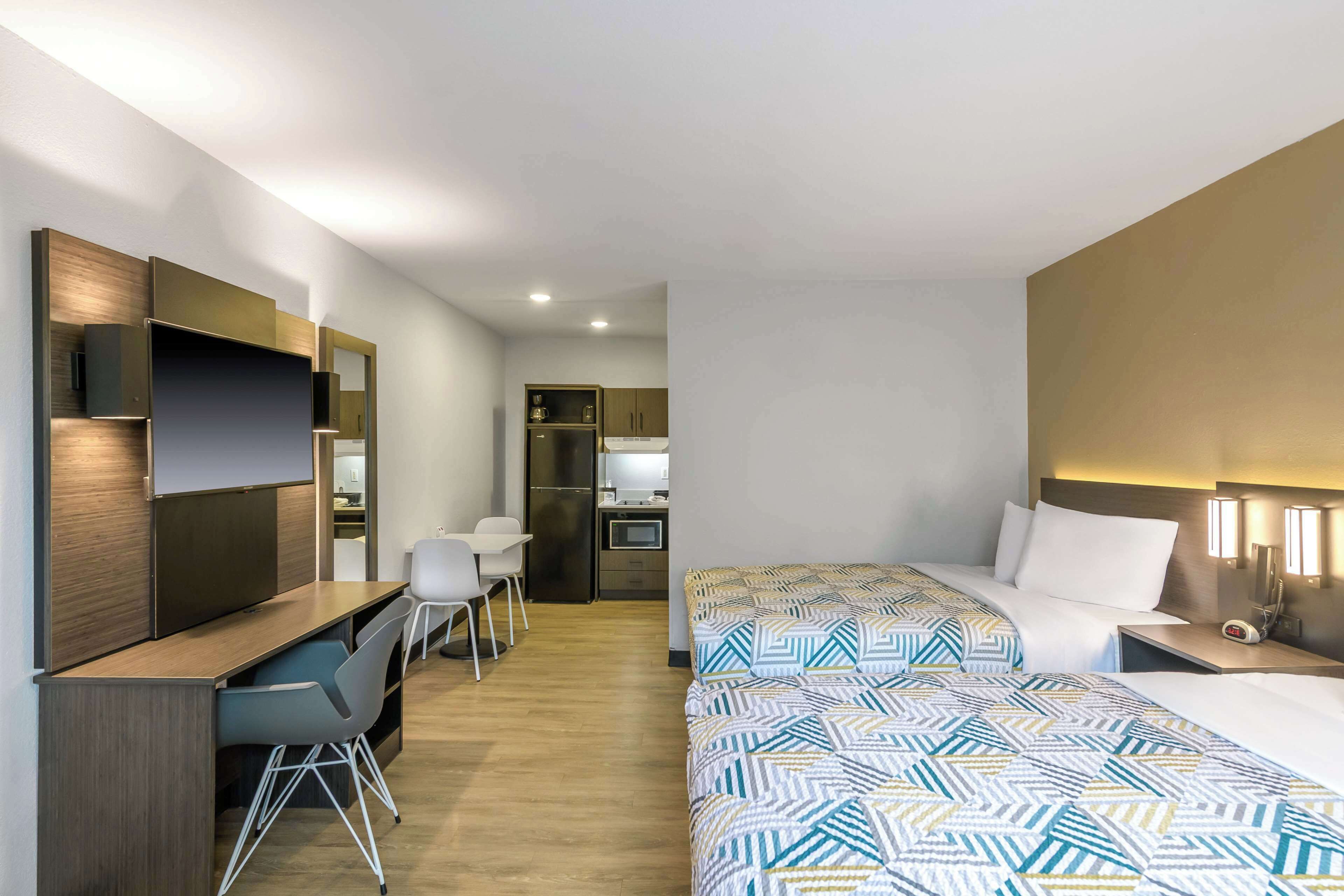 A hotel room with two beds, a desk, a chair, a kitchenette, and a mirror.