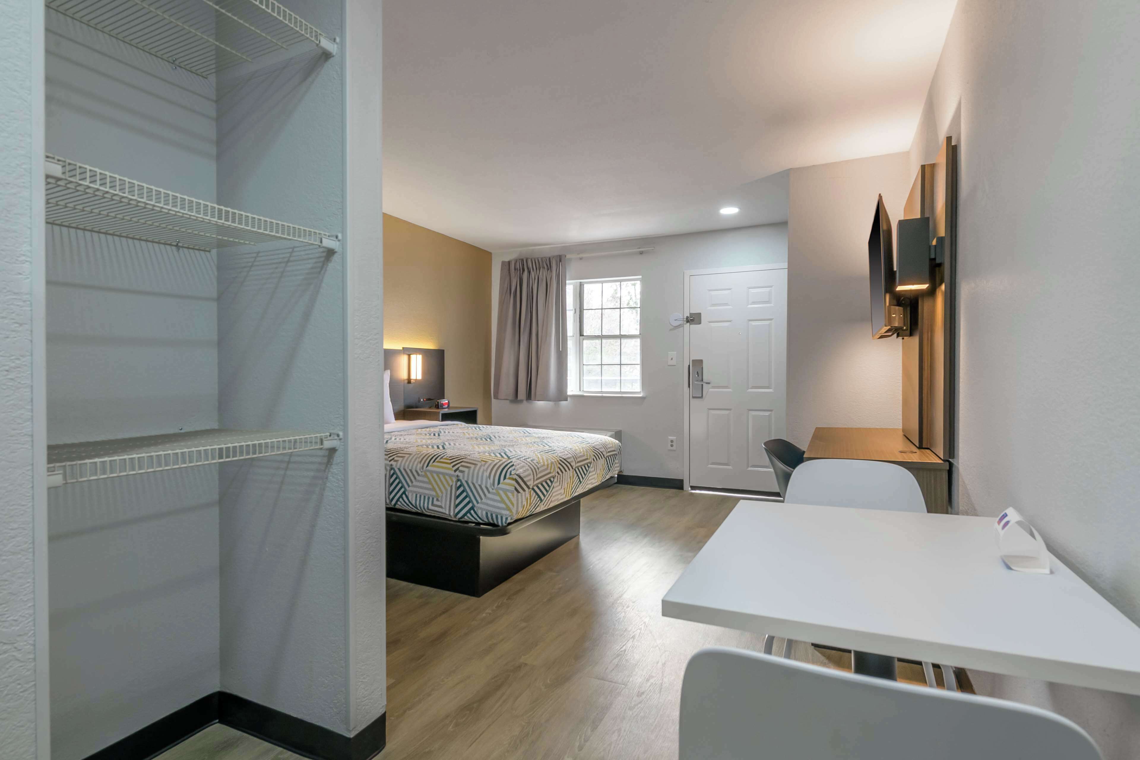 A modern hotel room with a bed, a desk, a chair, a TV mounted on the wall, and a window with curtains.