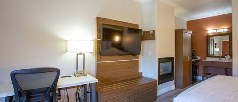 A hotel room with a bed, a desk with a chair, a television mounted on a wooden panel, a mirror above the vanity, and a fireplace.