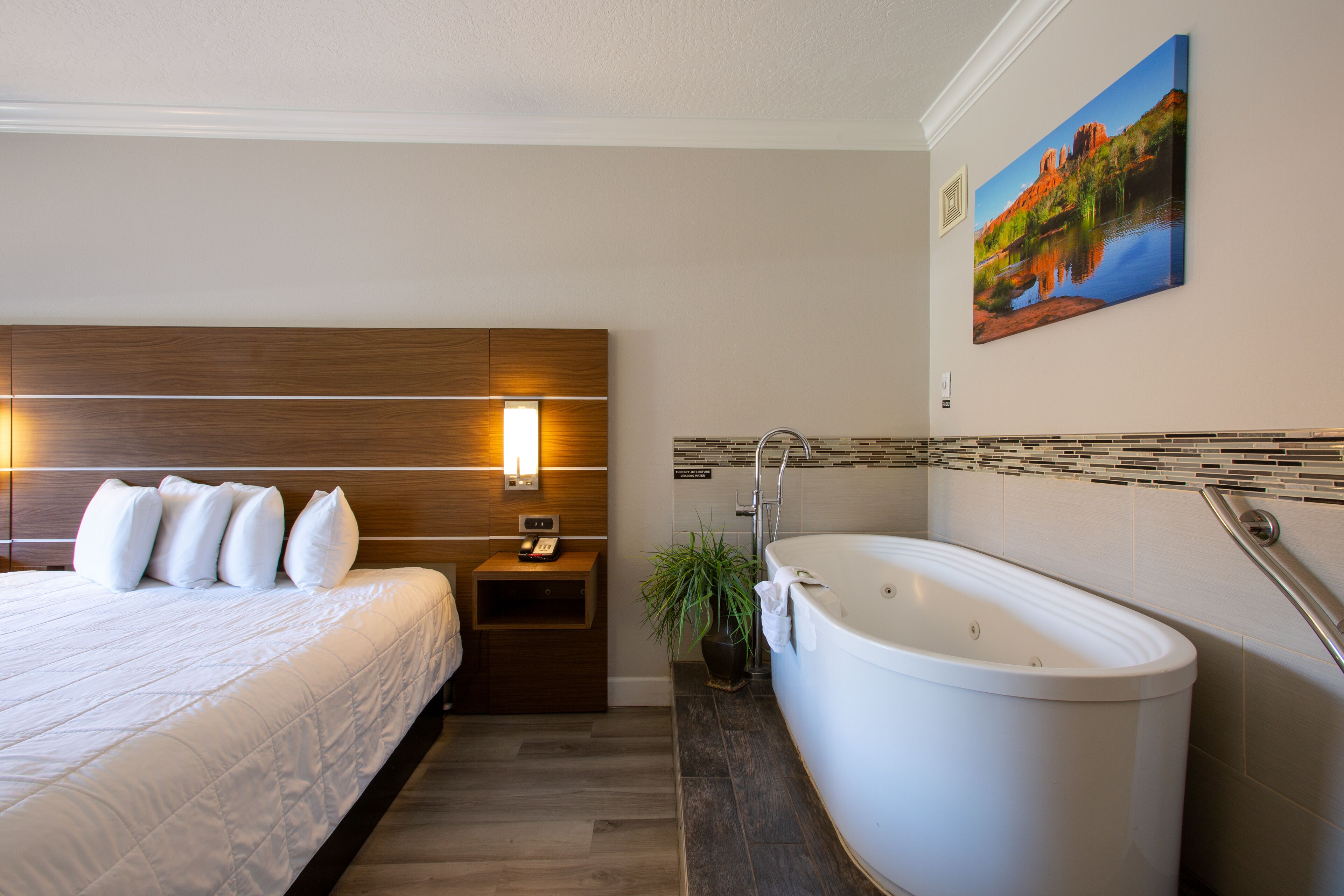 A hotel room with a large bed, a wooden headboard, a nightstand, a bedside table, a wall-mounted painting, and a freestanding bathtub.