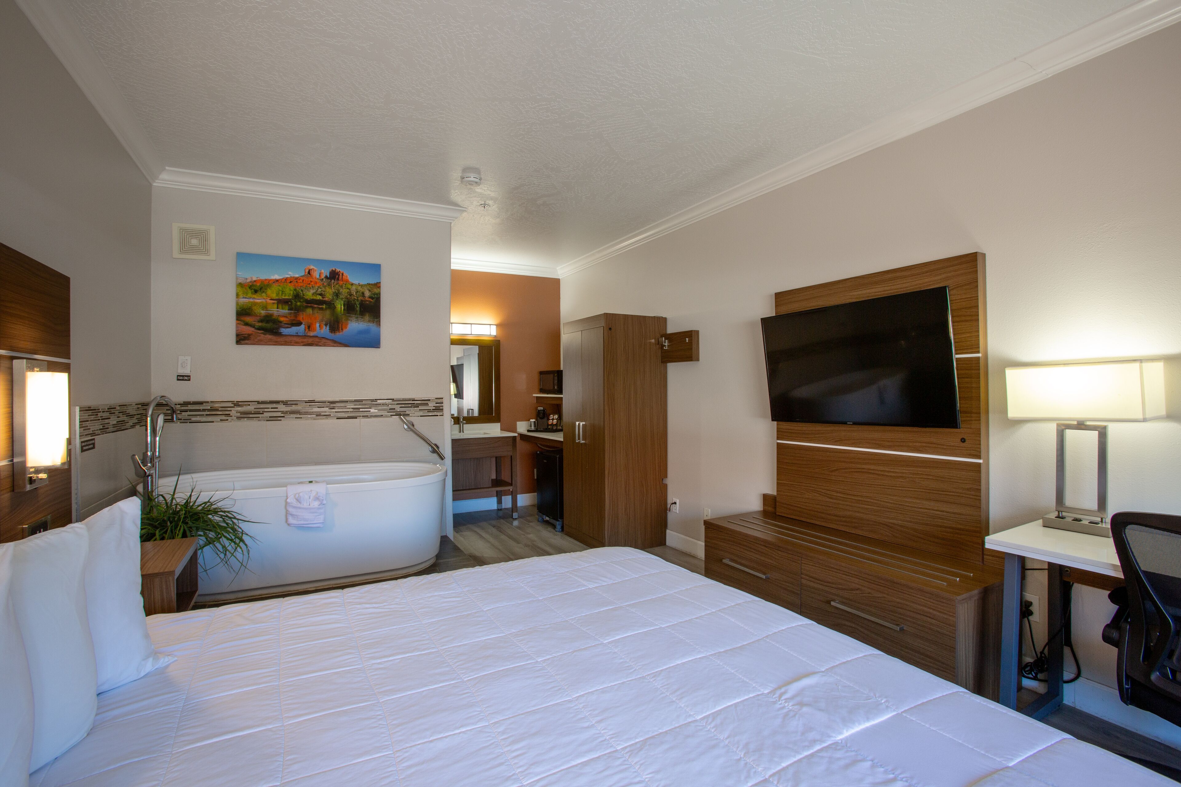 A hotel room with a large bed, a bathtub, a wooden wardrobe, a desk with a chair, and a TV mounted on the wall.
