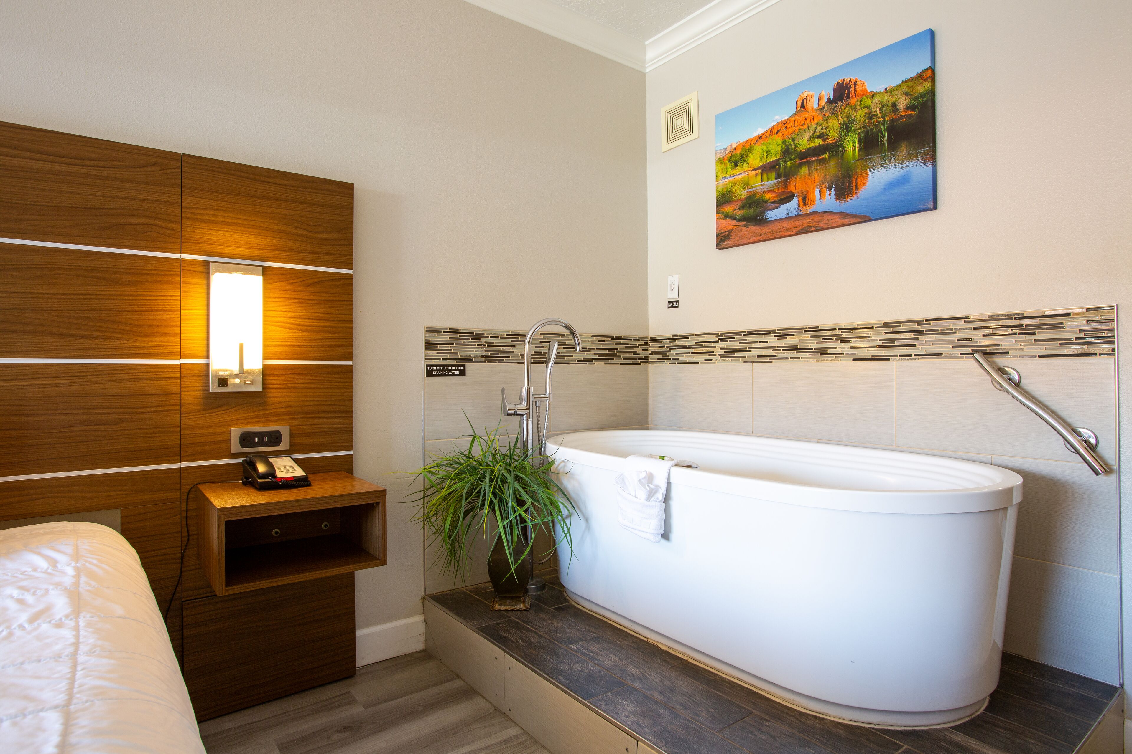 A modern bathroom with a large bathtub, a wooden vanity, a bedside table with a phone, and a potted plant.